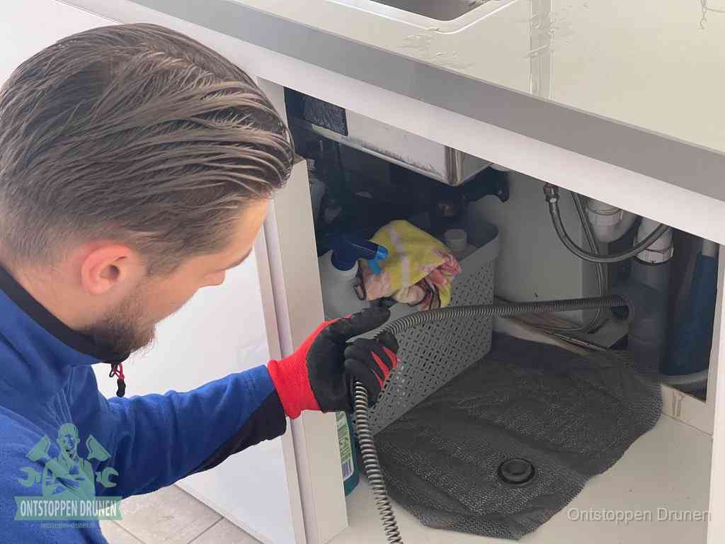 Een monteur voert een flexibele ontstoppingsveer in onder een keukenkastje om een verstopping voorbij de sifon te bereiken in Drunen