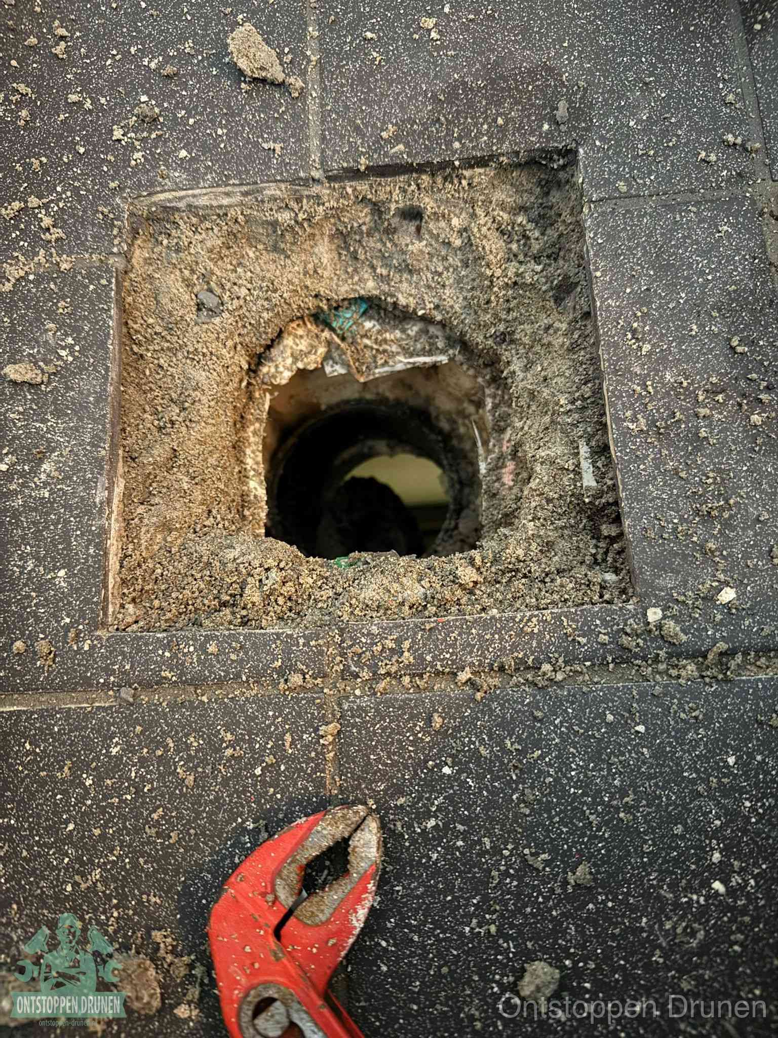 Een geopend rioolputje in een betontegelvloer met losgemaakt rooster en een zichtbare ronde afvoerbuis, het achterhalen waar de afvoerverstopping zit in Drunen