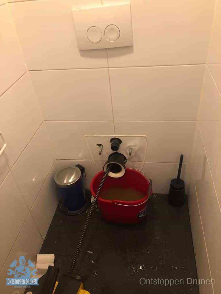 Een ontstopte wand-wc met verwijderde toiletpot waar een ontstoppingsveer in de wanduitgang verdwijnt, het achterhalen waar de verstopping zit in Drunen