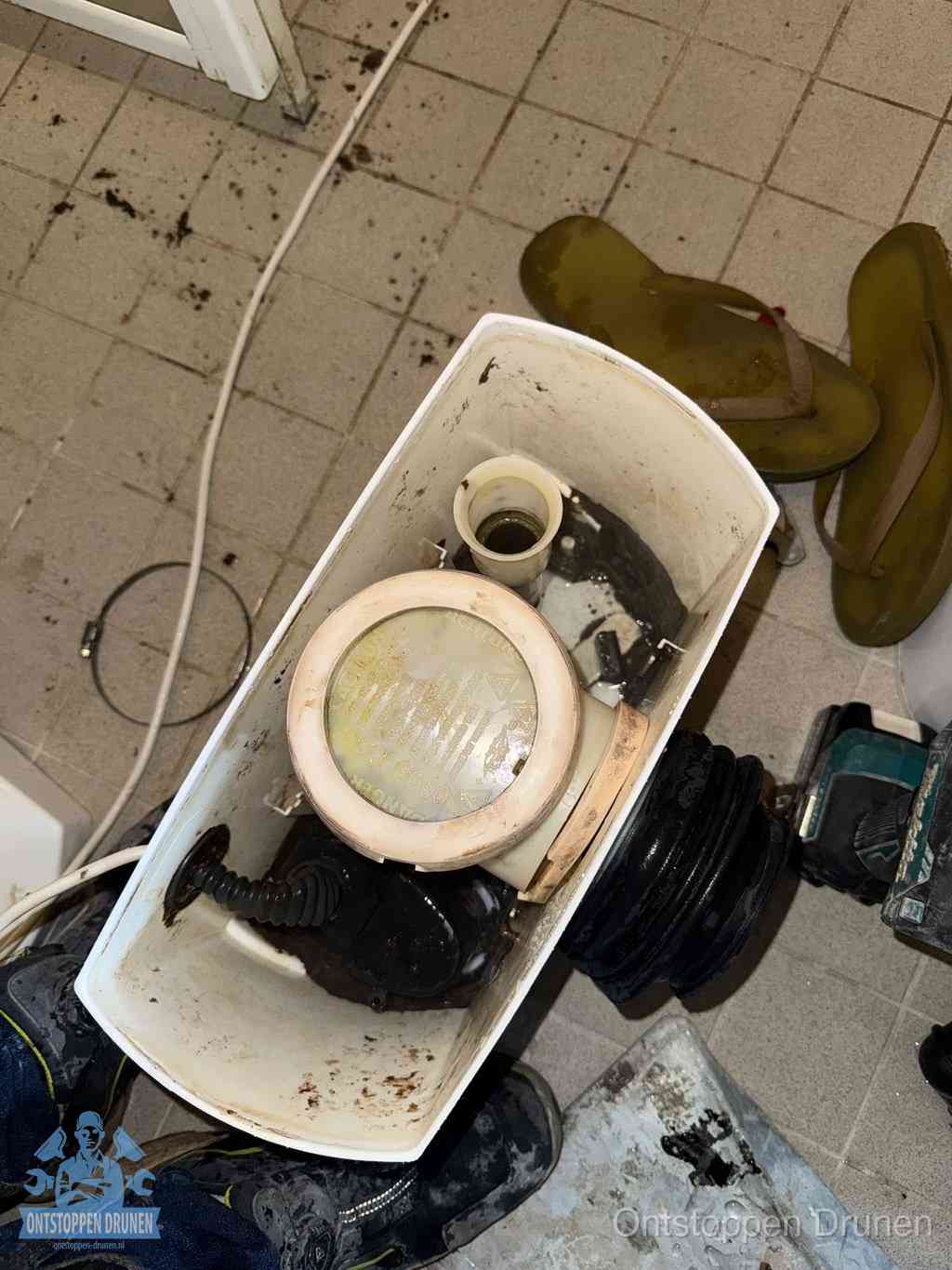 Een gedemonteerde toiletpot op de vloer met ontstoppingsveer en gereedschap ernaast, het verwijderen van een vastzittend voorwerp uit de wc in Drunen