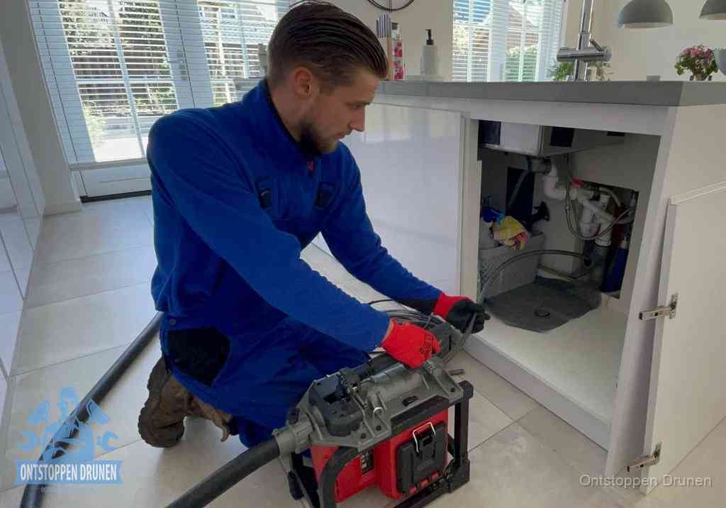 Een monteur voert een professionele elektrische ontstoppingsmachine in de afvoer onder de spoelbak tijdens het gootsteen ontstoppen in Drunen